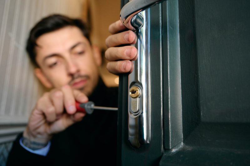 A dark haired man holding a door and screwdriver. The image is a close up of a silver handle and lock cylinder.