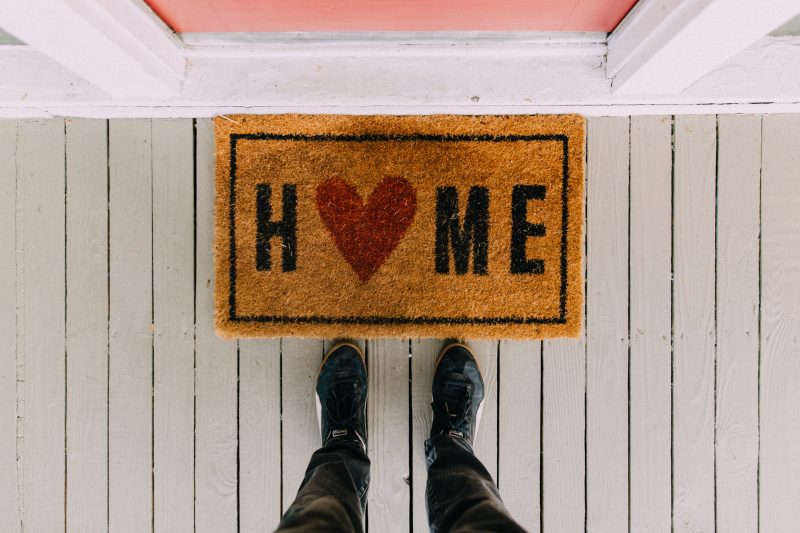 A man stands before a brown front door mat that says 'HOME'